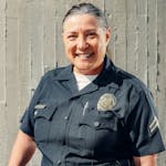 A cheerful policewoman smiling confidently in her uniform against a concrete wall.