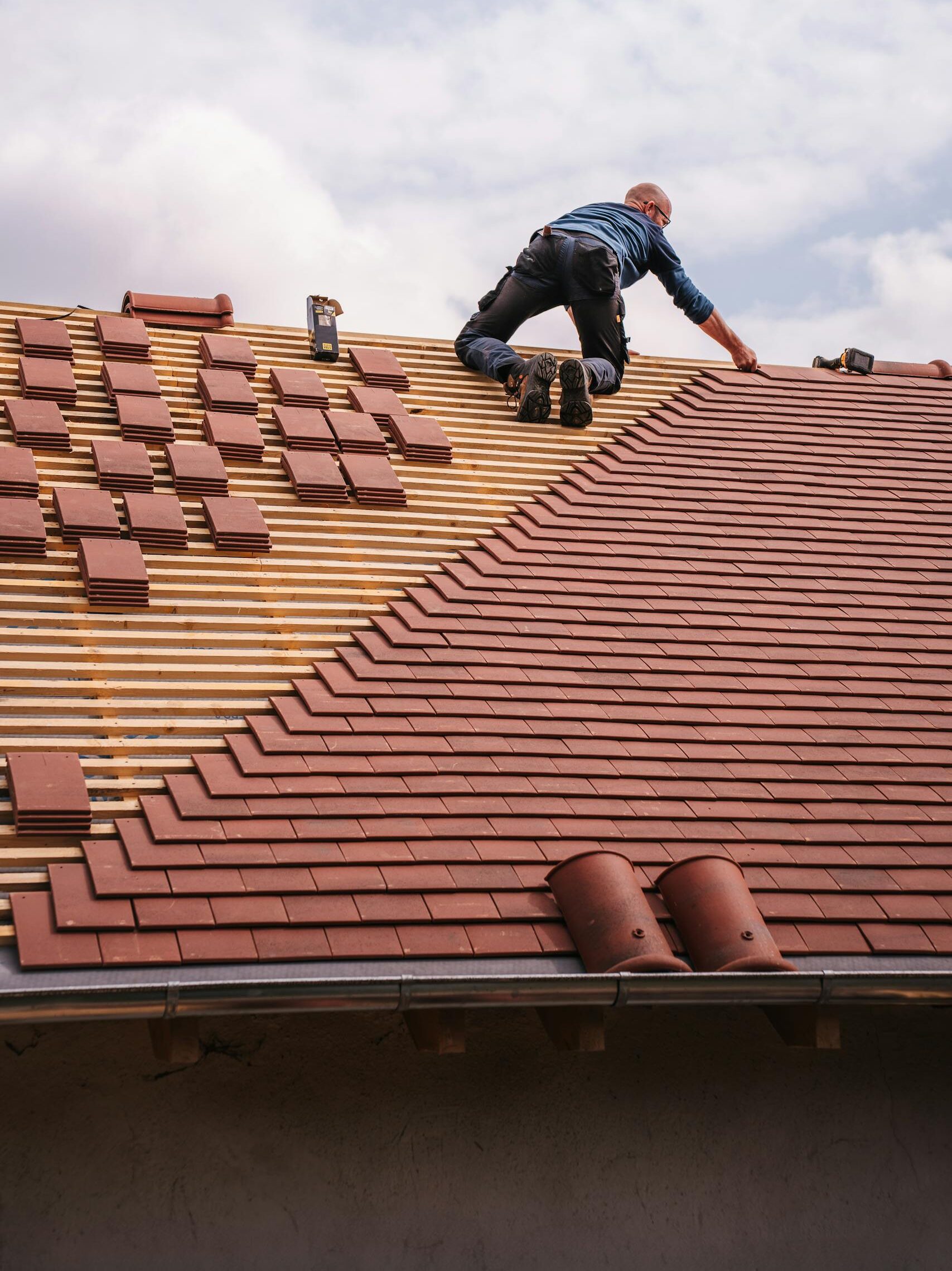 A roofer carefully installs shingles on a new residential roof under a cloudy sky.